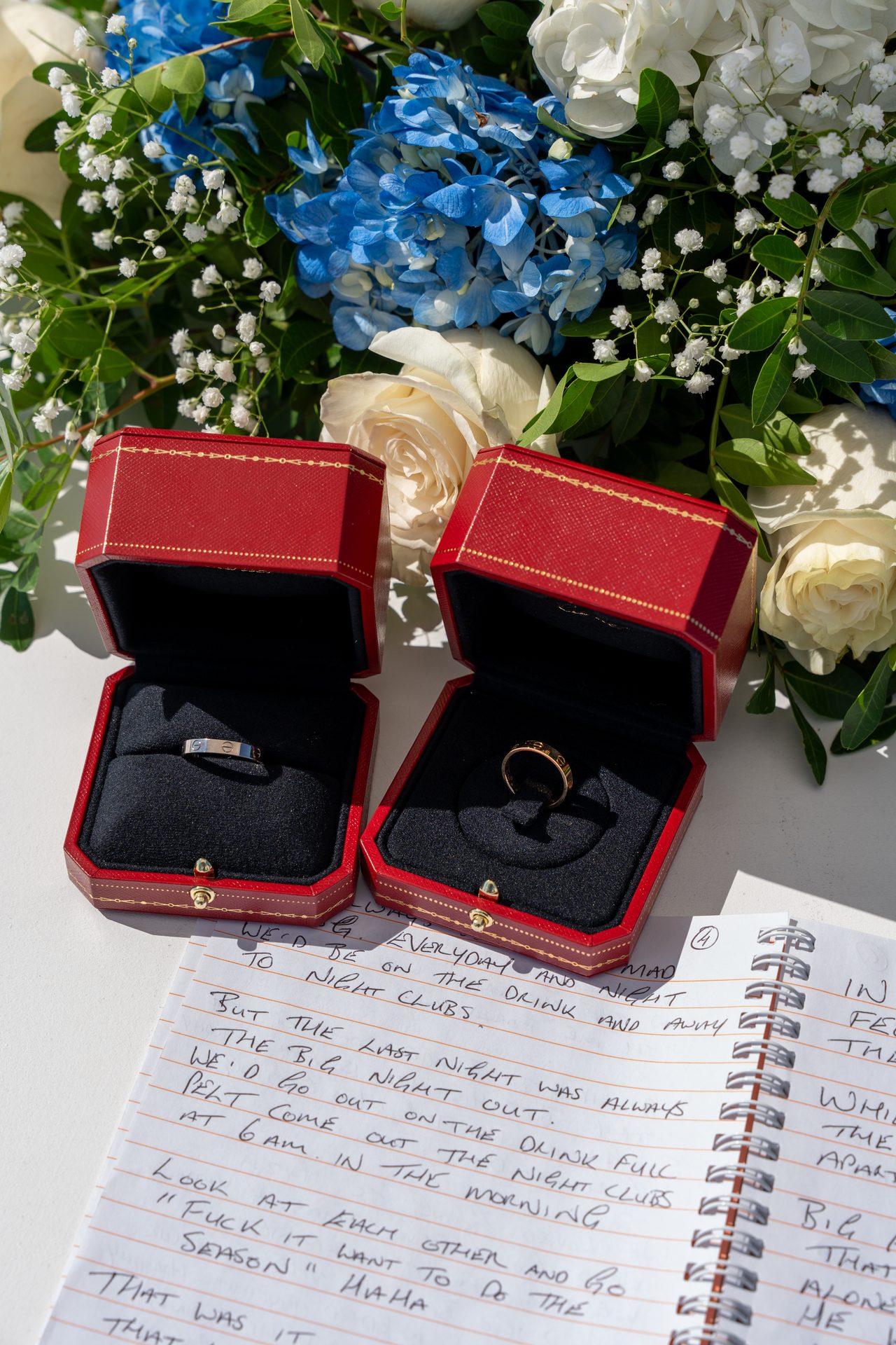 Wedding rings in red boxes beside handwritten vows and blue hydrangeas, Santorini wedding