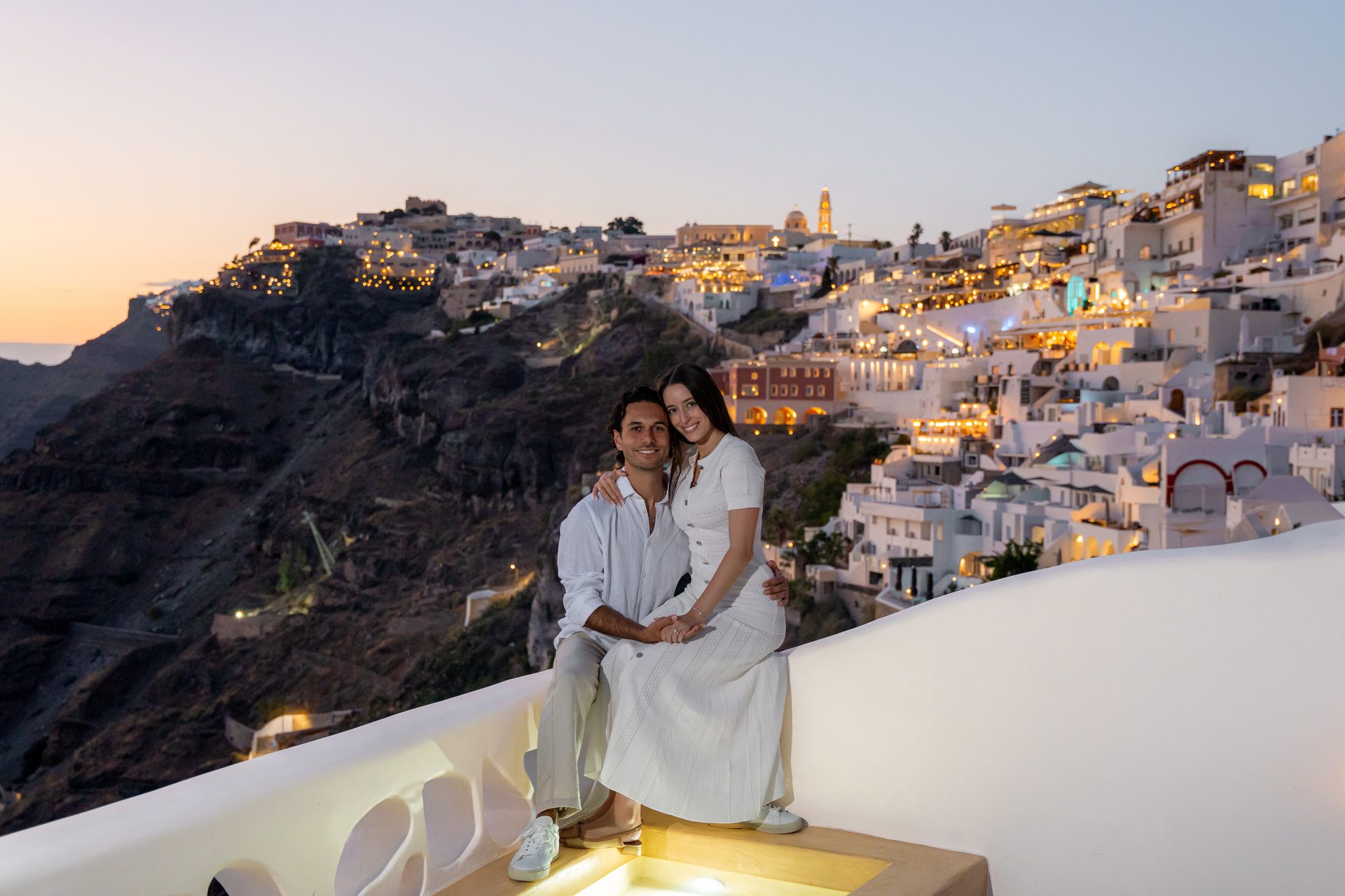 Engaged couple on a terrace above Fira at blue hour with the town lights glowing, Santorini