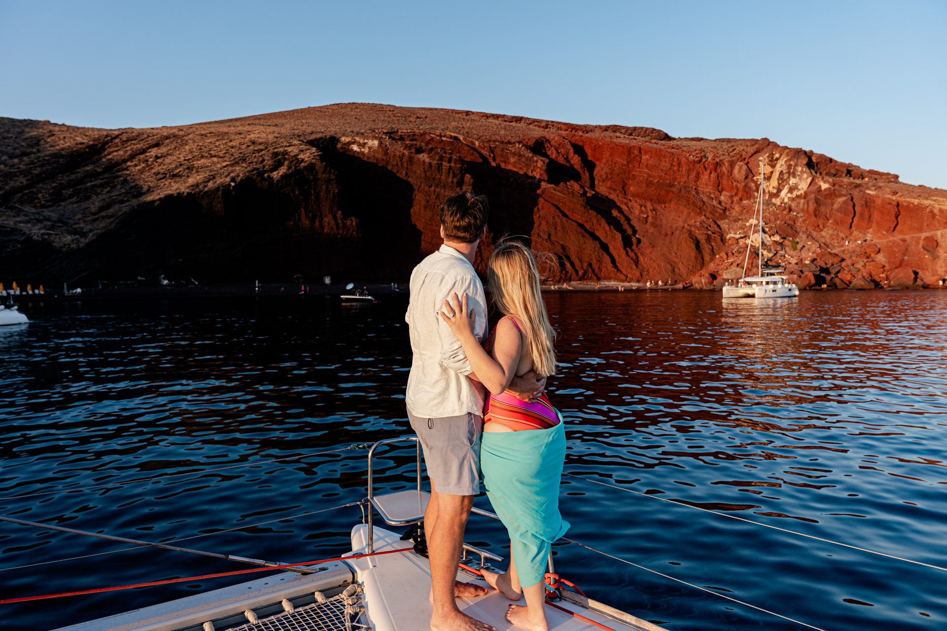 Couple on a catamaran bow beside the Red Beach cliffs at Akrotiri, Santorini
