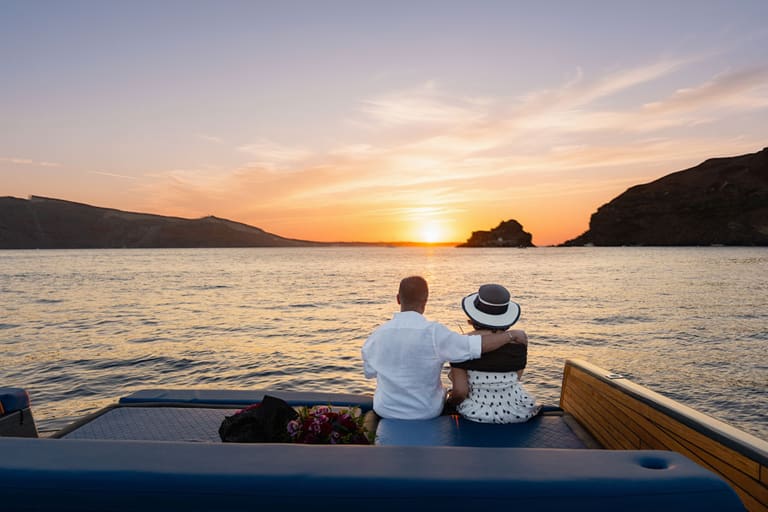 Couple sitting on the deck of a catamaran watching the sun set behind the Santorini caldera