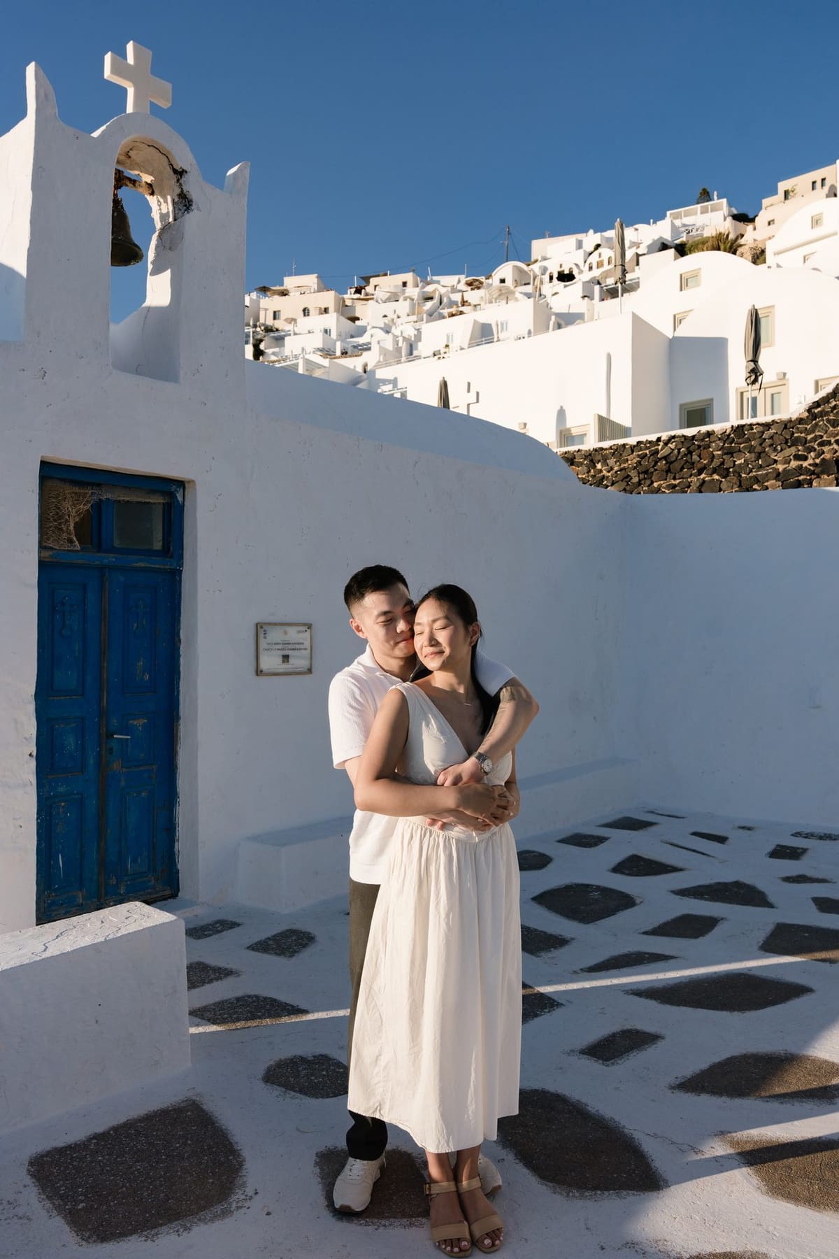 Couple in the white courtyard of an Oia chapel with a blue door, Santorini