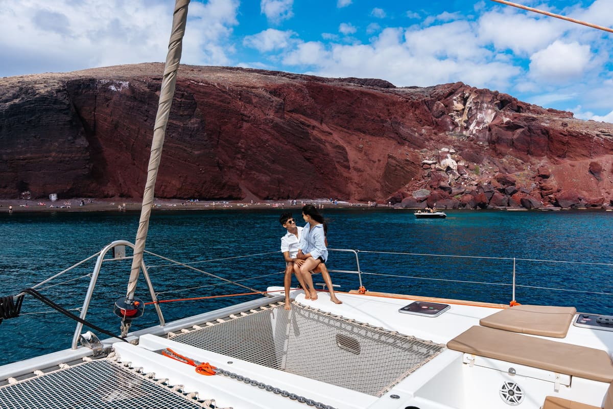 Couple seated on the bow of a catamaran below the Red Beach cliffs of Santorini