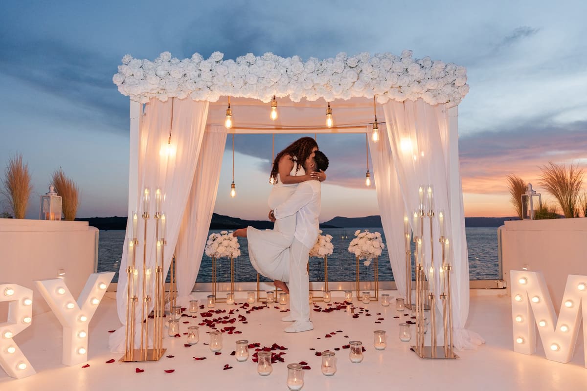 Man lifting his fiancée under the canopy beside lit MARRY ME letters at dusk, Santorini