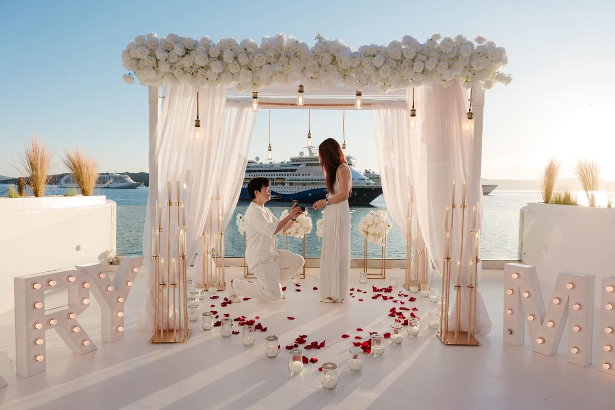 Man on one knee under a flower canopy with lit MARRY ME letters at a Santorini port venue