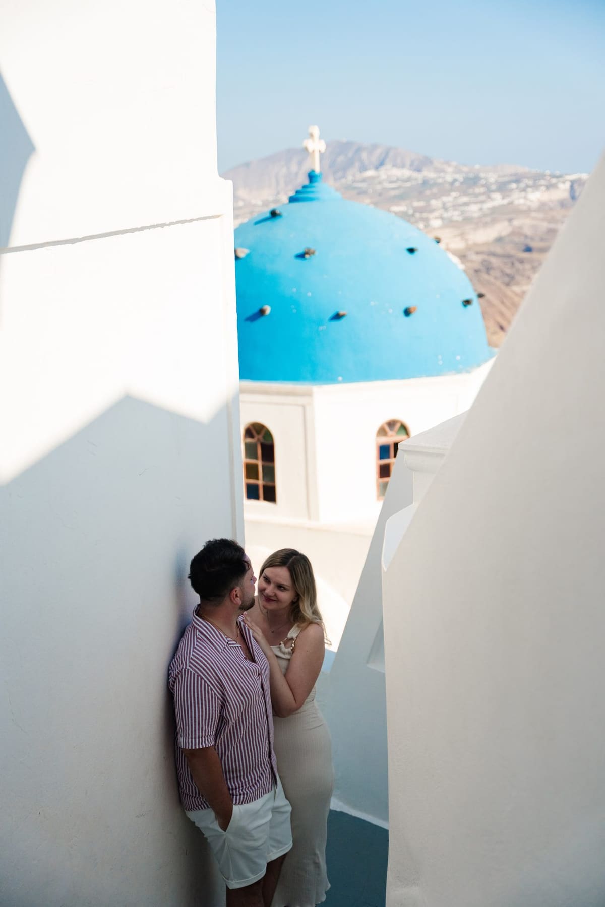 Couple leaning on a white wall beside a blue-domed church in Imerovigli, Santorini