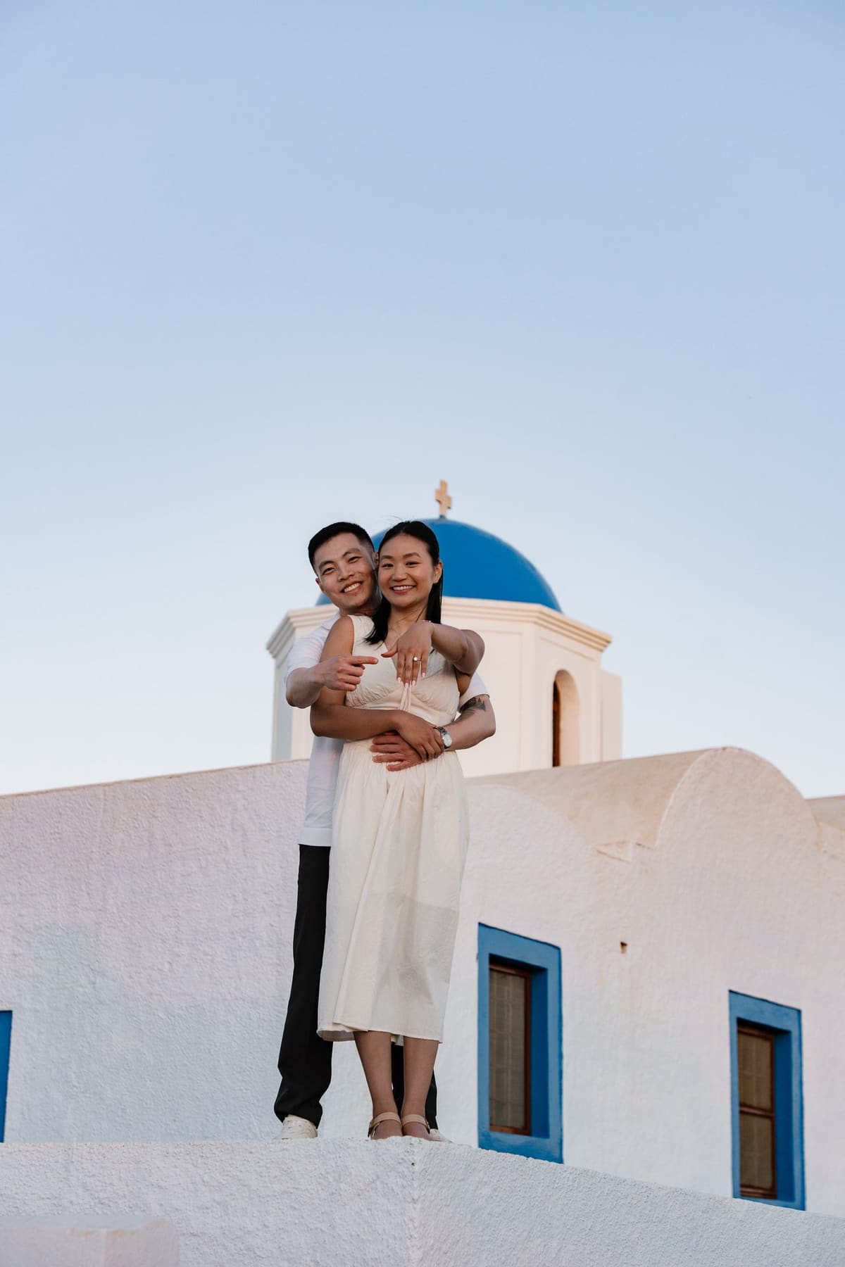 Couple embracing in front of a blue dome and white walls in Oia after their proposal