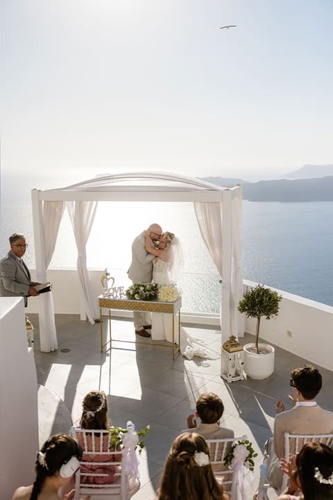 Wedding ceremony under a white gazebo on a caldera terrace with guests seated in the sun