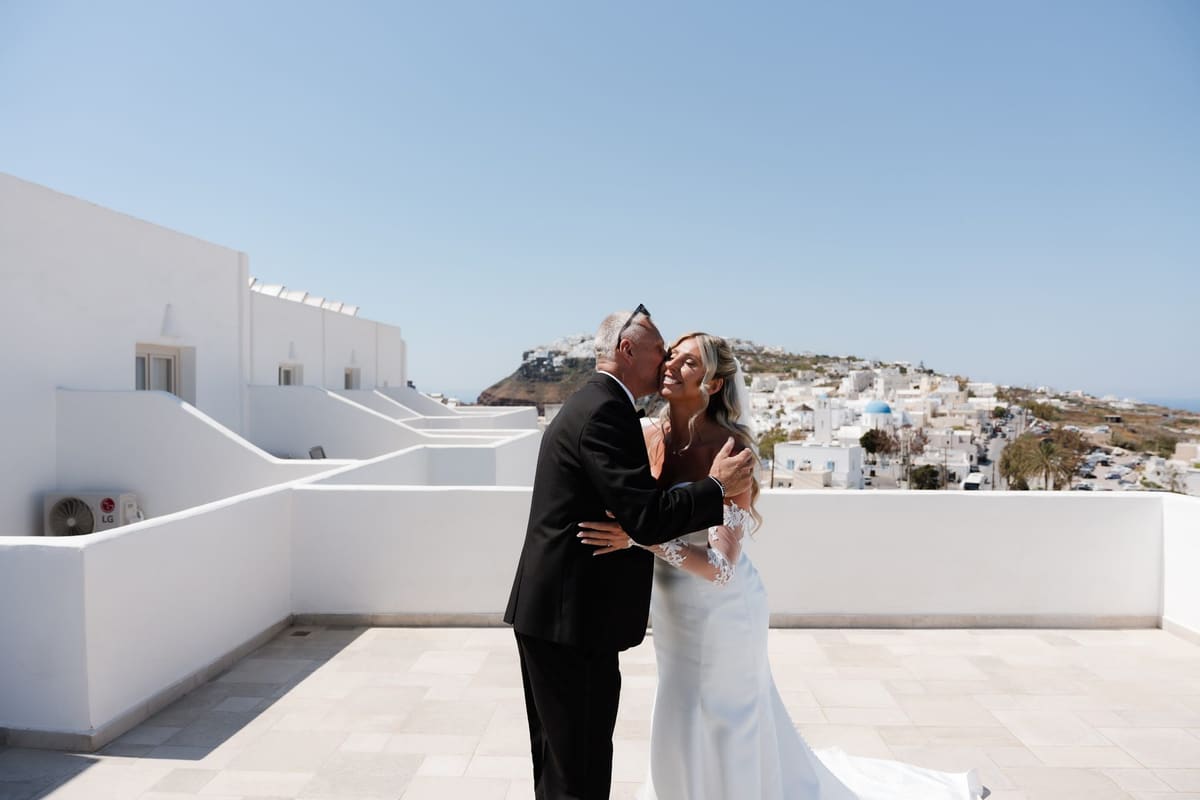 Groom in a black tuxedo embracing the bride on a white rooftop above Fira, Santorini