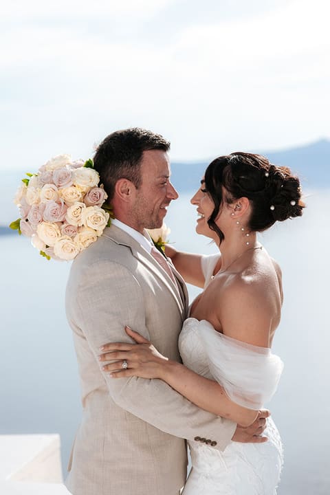 Bride with a white bouquet and groom in a beige suit embracing above the caldera, Imerovigli