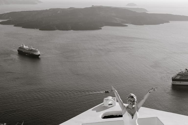 Bride cheering with open arms on a caldera terrace with cruise ships below, black and white