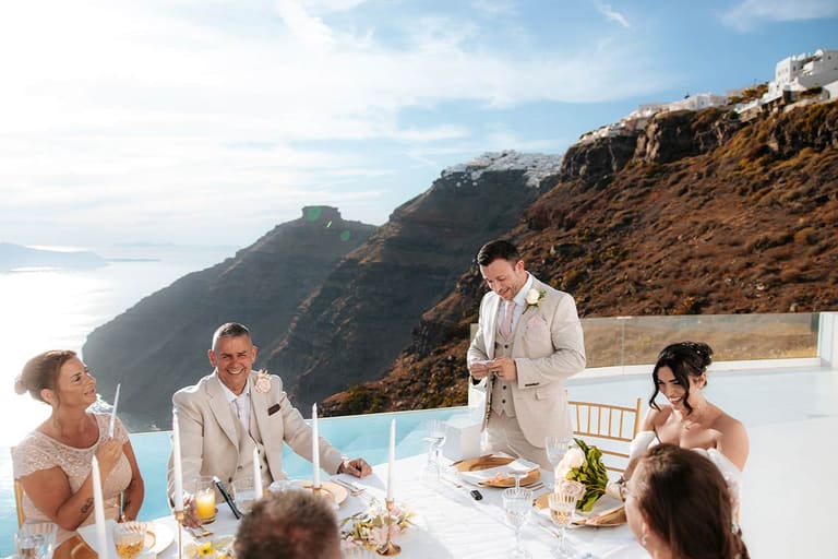 Wedding guests at a long dinner table above the Santorini caldera in Imerovigli at golden hour