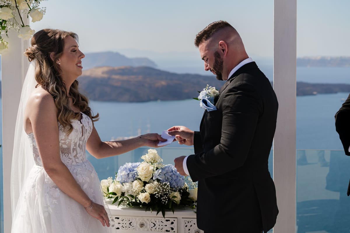 Bride in lace placing the ring on the groom's finger with the caldera behind, Santorini