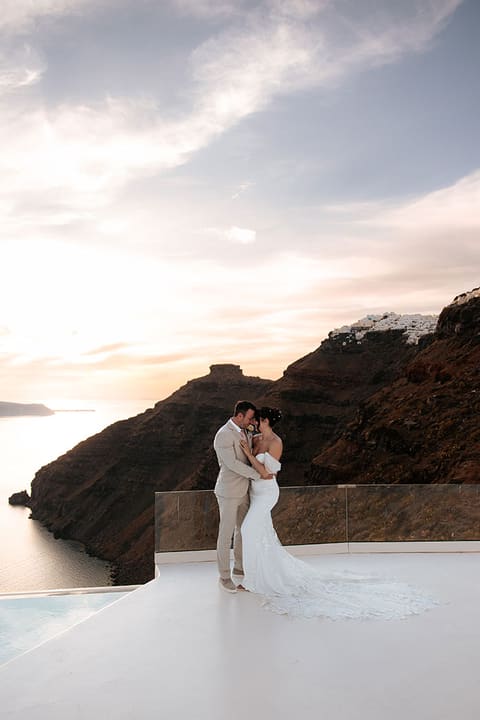 Bride and groom embracing on a caldera terrace in Imerovigli at sunset, Skaros Rock behind them