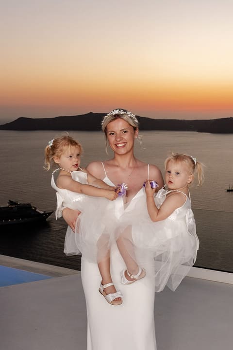 Bride holding two toddlers on a caldera terrace at sunset, Santorini family wedding moment