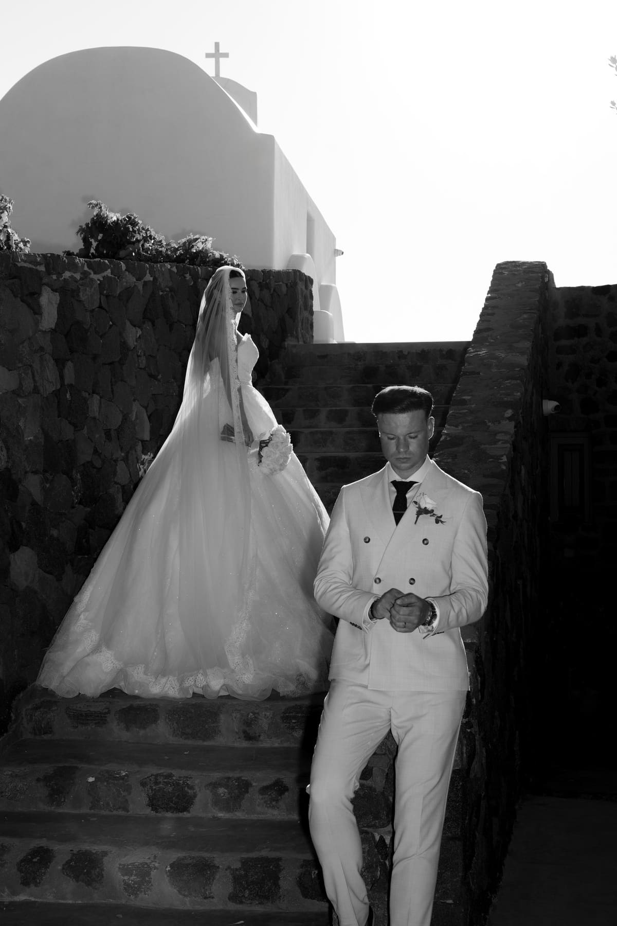 Bride and groom in white descending the stone steps of a Santorini chapel, backlit